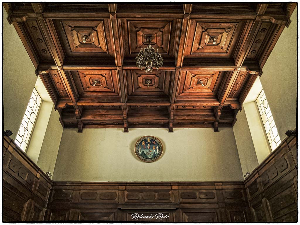 Artesonado de madera del Salón General Mayor “Adolfo Mijangos López”, cubriendo el techo y zócalo del salón, adornado con una lámpara de cristal colgante y el escudo de la USAC al fondo.